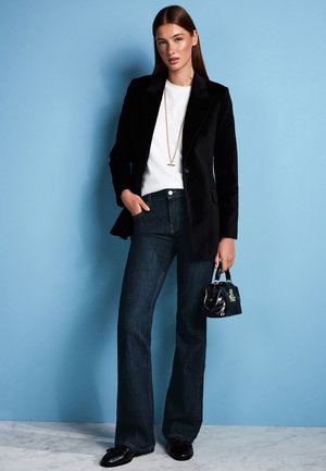 Kvinde står mod en blå væg iført sort blazer, hvid top, mørke jeans, sorte sko, gyldent halskæde og holder en lille sort håndtaske.