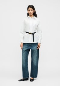 Λευκό πουκάμισο με κουμπιά, μακρύ μανίκι και τσέπη στο στήθος, σφιγμένο στη μέση με μαύρη ζώνη. Συνδυασμένο με φαρδιά μπλε τζιν παντελόνια.