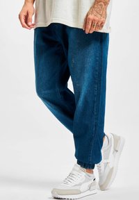 Pantalones jogger de mezclilla en azul oscuro con un ajuste relajado, con puños elásticos y sutiles detalles desgastados, combinados con deportivas blancas y grises.