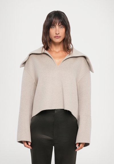 Beige cropped sweater med en stor foldet krave, brede ærmer og en blød, tekstureret overflade. Matchende med sorte, strømlinede bukser.