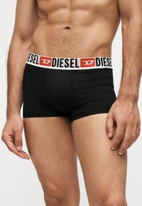 Άνδρας κορμός που φοράει μαύρα μπόξερ της Diesel με λευκή μέση με κόκκινο και μαύρο λογότυπο Diesel, που στέκεται σε απλό φόντο.