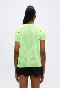 Γυναίκα που φοράει κοντομάνικη μπλούζα από διαφανές δίχτυ σε νέον πράσινο και μαύρο σορτς, στέκεται με την πλάτη στην κάμερα μπροστά σε απλό φόντο.