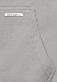 Tasca in tessuto grigio con cuciture angolari e un piccolo tag metallico rettangolare con la scritta "bazix republiq" cucito nell'angolo in alto a sinistra.