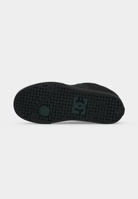 Semelle d'une chaussure de sport noire montrant un motif de bande de roulement avec des formes rondes et rectangulaires et un logo au centre sur un fond uni.