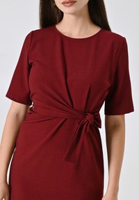 Robe bordeaux en tissu lisse, avec un col rond, des manches courtes et un détail de nœud à l'avant à la taille.