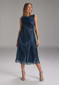 Femme souriante portant une robe midi sans manches bleu scintillant et des talons à bride de cheville bleus, se tenant devant un fond gris uni, regardant sur le côté.