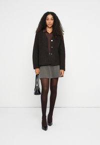Vrouw in een bruine knopenka cardigan, bruine shirt, grijze rok, donkere panty, zwarte hakken, met een zwarte handtas, staand tegen een witte achtergrond.