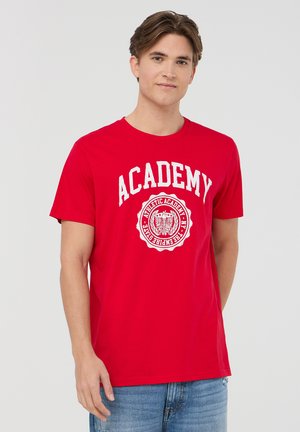 T-shirt rossa a maniche corte in cotone, con testo bianco "ACADEMY" e un grafiche logo circolare sul davanti. Taglio dritto.