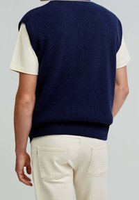 Granatowy szydełkowany kamizelka bez rękawów, noszona na jasnym kremowym t-shircie, w połączeniu z jasnymi spodniami z tylnymi kieszeniami.