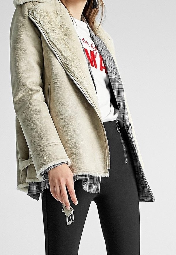 Beige Shearling-Jacke mit Kapuze, ausgestattet mit einem Reißverschluss und einer Karolining. Getragen über einem weißen Hemd und schwarzen Hosen, mit einem Schlüsselanhänger.