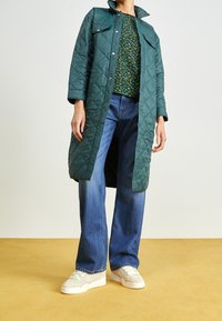 Abrigo largo acolchado verde, top con estampado floral, jeans de pierna ancha azules y zapatillas crema. Los botones y los detalles del cuello realzan el abrigo.