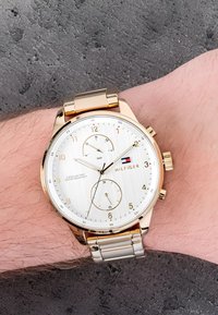 Złoty zegarek chronograficzny z białą tarczą, trzema subzegarami i wzorzystą skórzaną bransoletą na nadgarstku, na tle o fakturze.