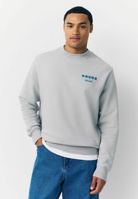 Šedý crewneck svetr vyrobený z bavlny, s modrou grafickou potiskem na hrudi. Žebrované manžety a lem, uvolněný střih.