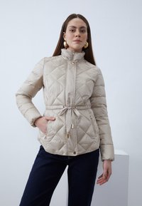 Quilted beige pufferjakke med høy krage, justerbart snøring i midjen og to lommer, kombinert med mørke jeans.