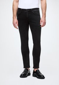 Feszes szabású fekete farmernadrág klasszikus öt zsebes dizájnnal, övhurkokkal és diszkrét varrási részletekkel, fekete cipővel párosítva.