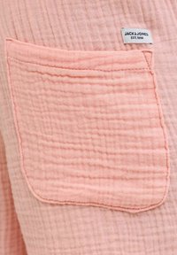 Ροζ υφασμάτινη τσέπη με ορατές ραφές και μια μικρή λευκή ετικέτα "JACK & JONES EST. 1990" ραμμένη κοντά στην άνω άκρη.