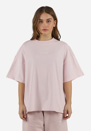 Fiatal nő, aki túlméretezett világos rózsaszín pólót és hozzáillő rövidnadrágot visel, előre néz, semleges arckifejezéssel.