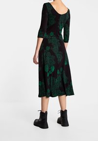 Robe noire à motif floral vert, manches trois-quarts et dos ouvert, associée à des bottines noires. Texture lisse et longueur mi-mollet.