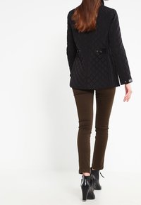 Veste noire matelassée au design ajusté, avec deux poches latérales, des détails zippés et des épaules structurées. Associée à un pantalon ajusté marron.