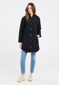 Giacca lunga nera trapuntata con bottoni a pressione e tasche laterali. Indossata con jeans azzurri e sneakers beige. Tessuto morbido, design semplice.