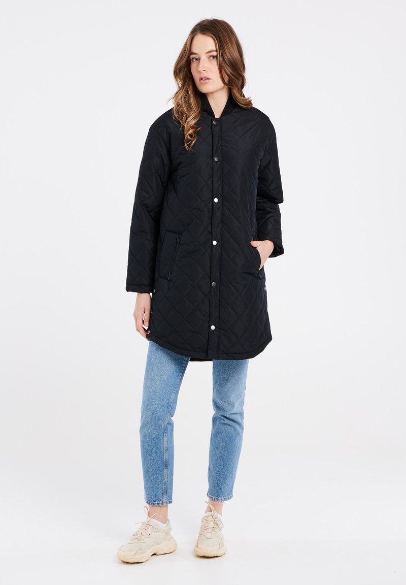 Giacca lunga nera trapuntata con bottoni a pressione e tasche laterali. Indossata con jeans azzurri e sneakers beige. Tessuto morbido, design semplice.