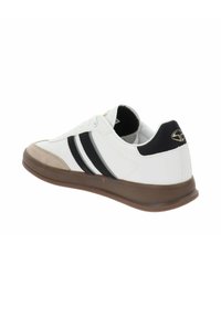 Zapatilla deportiva blanca con rayas negras y puntera de ante color tostado. Cuenta con una suela de goma y un diseño de caña baja. Logo dorado en el talón.