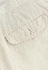 Una tela de color beige claro presenta una superficie texturizada con un bolsillo de solapa y un botón blanco. Las costuras son visibles a lo largo de los bordes del bolsillo.