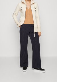 Hoodie zippé beige clair avec cordons, cardigan à côtes orange, et pantalon large à rayures bleu marine. Baskets noires avec semelle blanche.