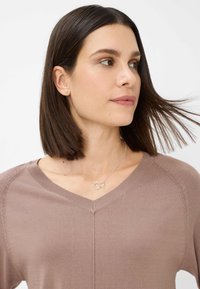 Beige V-hals sweater med strikstruktur, der har diskrete detaljer i siderne. Båret med en sølvhalskæde med to indviklede cirkler.