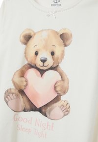 Top in cotone bianco per pigiama con un'illustrazione di un orso che tiene un cuore rosa. Il testo sottostante recita "Buona Notte, Dormi Bene." Decorato con un fiocco.