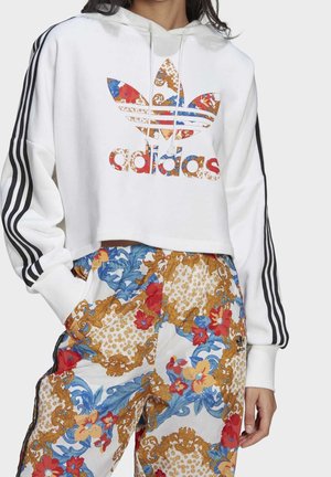 Kobieta w białej skróconej bluzie z kapturem Adidas z kwiatowym logo i paskowanymi rękawami, w połączeniu z wielokolorowymi spodniami w kwiatowy wzór.
