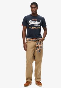 Marineblauwe katoenen t-shirt met witte en gele grafische tekst, beige katoenen broeken, geruite flanellen stof om de taille geknoopt, bruine schoenen.