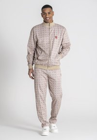 Lichtroze trainingspak gemaakt van zachte stof, met een subtiel logo-patroon, een ritsjack en bijpassende broek. Met crèmekleurige manchetten.