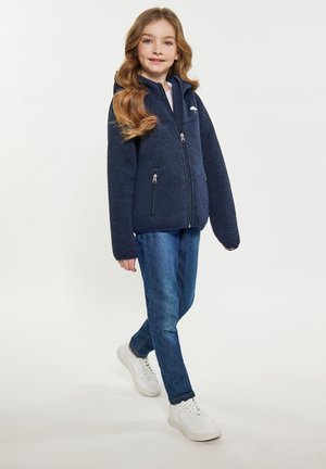 Giacca zip-up blu navy con cappuccio, dotata di due tasche con zip e finitura testurizzata, abbinata a jeans blu e sneaker bianche.