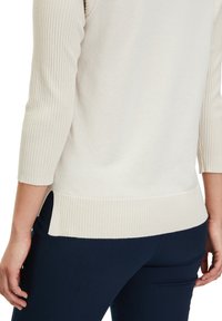 Helle beige gerippte Strickpullover mit 3/4-Ärmeln, seitlichen Schlitzen und einem kontrastierenden, strukturierten Saum. Kombiniert mit marineblauen Hosen.