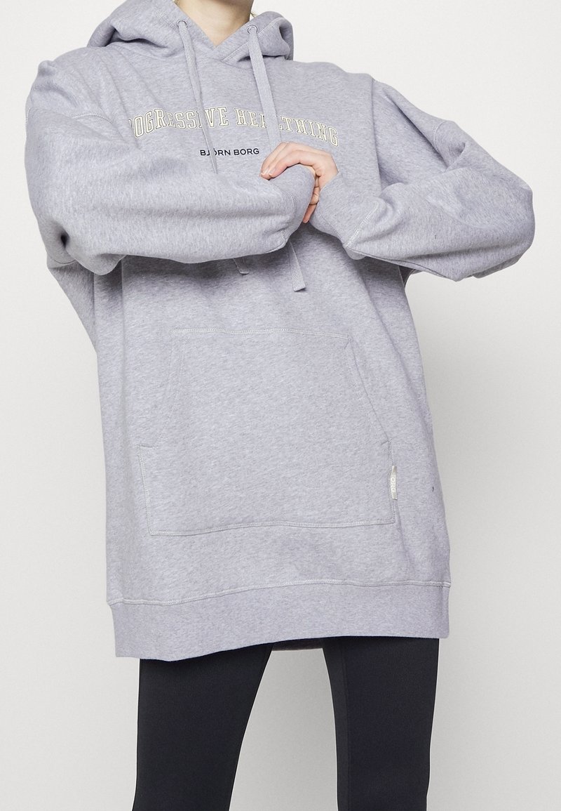 Sweat à capuche gris clair avec cordon de serrage, poche avant et texte "PROGRESSIVE HEALTHING BJORN BORG" en jaune et noir. Matière en coton, coupe décontractée.