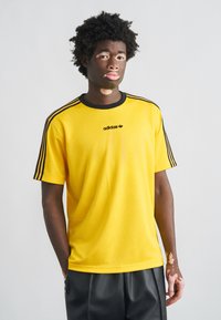 Κίτρινη μπλούζα Adidas με μαύρες ρίγες στους ώμους, λαιμόκοψη τύπου crew, και μικρό λογότυπο στο στήθος, φτιαγμένη από απαλό ύφασμα.