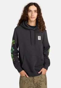 Jongere met lang krullend haar, gekleed in een donkere hoodie met groene botanische ontwerpen op de mouwen en een patroon van olijfgroene broek.