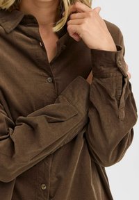 Camisa de pana marrón con cierre de botones en la parte delantera, grandes puños y una tela texturizada con rayas verticales.