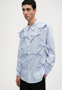 Camisa xadrez azul clara com vários laços de tecido nos ombros e mangas, frente com botões e colarinho clássico.