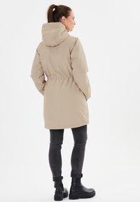 Beige parka dengan tudung, pinggang yang diketatkan, dan lengan panjang, dipadukan dengan celana jeans abu-abu gelap dan sepatu bot pergelangan hitam, dilihat dari belakang.