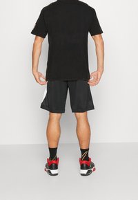 Hombre de pie de espaldas, vistiendo una camiseta negra, pantalones cortos negros con paneles blancos, calcetines negros y zapatillas negras y rojas.