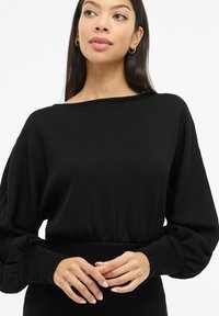 Musta ribbineulottu pitkähiha-paita, jossa on leveä pääntie ja pullotetut hihat, istuva vyötärö ja teksturoitu kangas. Hopeiset sormukset käsissä.