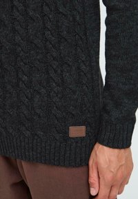 Mørk grå kabelstrikket genser med et teksturert mønster og ribbede mansjetter, med et brunt skinnlogo merke i bunnen.