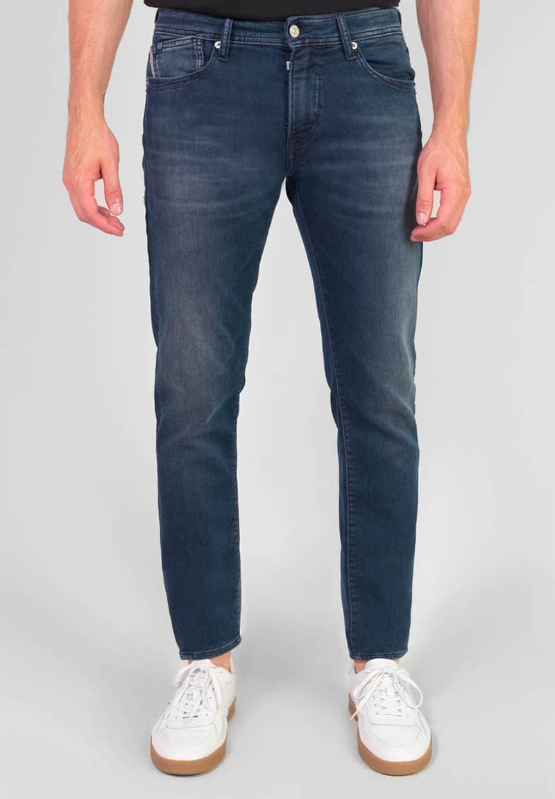 Jeans de denim azul oscuro con un ajuste slim, cinco bolsillos y costuras en contraste. Combinados con zapatillas blancas con suela marrón.