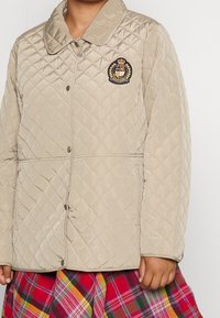 Beige kviltad jacka med en slät yta, knappstängning och en marinblå emblem föreställande en krona och lagerkrans, i kombination med en rutmönstrad kjol.