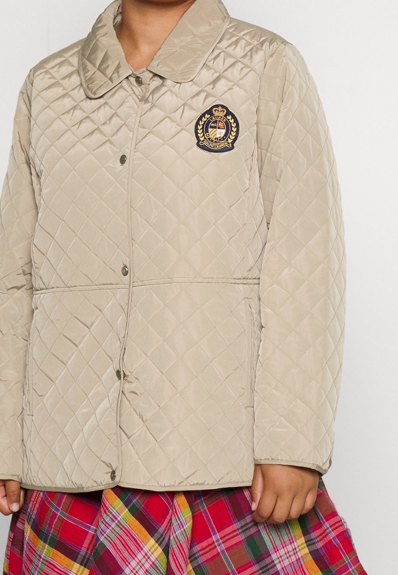 Beige kviltad jacka med en slät yta, knappstängning och en marinblå emblem föreställande en krona och lagerkrans, i kombination med en rutmönstrad kjol.