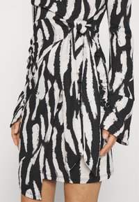 Šaty se zebřími pruhy v černobílé barvě, s dlouhými rukávy, uzlovým detailem v pase a strukturovanou tkaninou. Obsahují vrstvený design a rozšířené manžety.