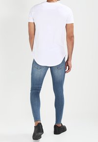 Man som står med ryggen mot kameran, iklädd en vit kortärmad skjorta, blå slim fit-jeans och svarta sneakers mot en enfärgad ljus bakgrund.