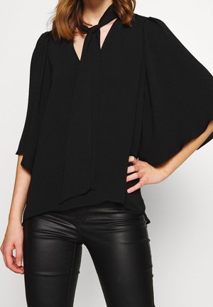 Femme portant un chemisier noir ample avec un col noué et des manches larges, associé à un pantalon en cuir noir ajusté, main sur la hanche.